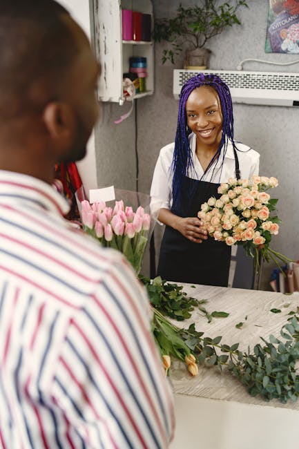 A young woman with a bright smile holds a bouquet of light pink roses with lush green foliage in a floral workshop, standing behind a white tabletop. She is wearing a white top and black apron, with l