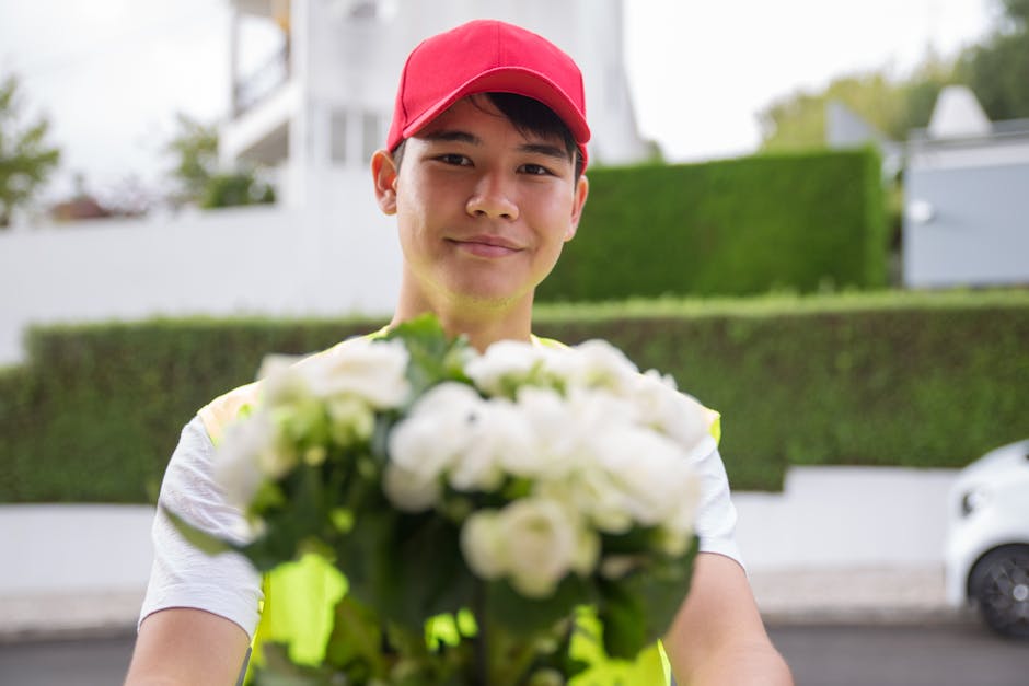 A smiling young male flower delivery courier wearing a red cap and a high-visibility vest holds out a bouquet of fresh white flowers towards the camera. The bouquet features a mix of large white bloom