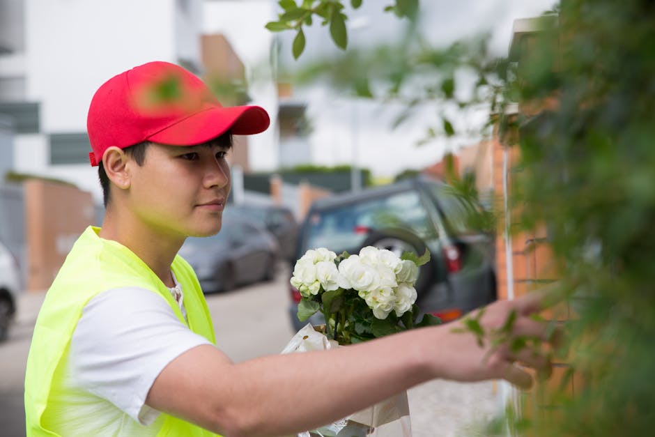 A young male florist, wearing a red cap and a neon yellow vest over a white t-shirt, is arranging a floral bouquet outdoors. He holds a bunch of white hydrangeas with green leaves in one hand while ca